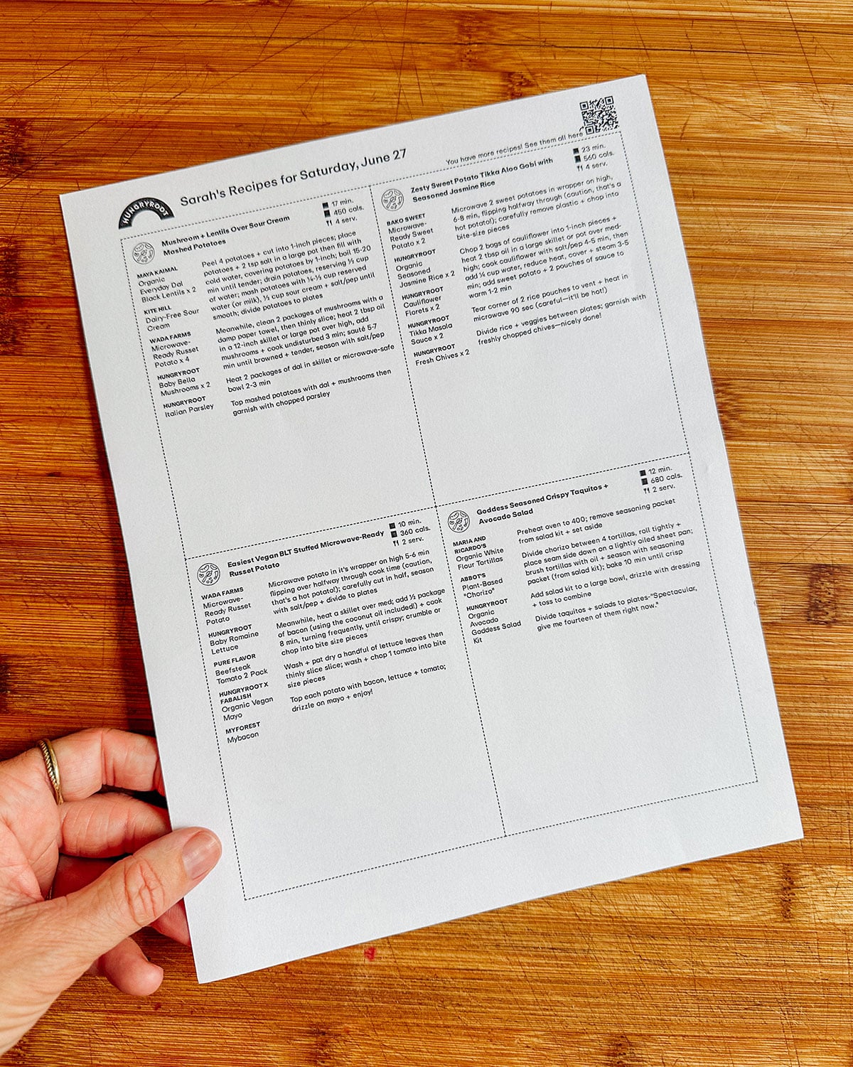 A hand holds a printed recipe sheet with four recipes and titles on a wooden surface. The sheet is labeled “Sarah’s Recipes for Saturday, June 27.”.