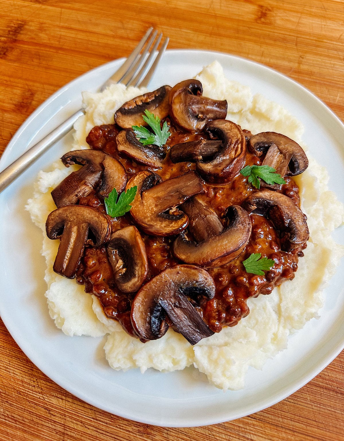 Plate of mashed potatoes topped with lentil stew, sautéed mushrooms, and parsley, with a fork on the side, on a wooden surface.