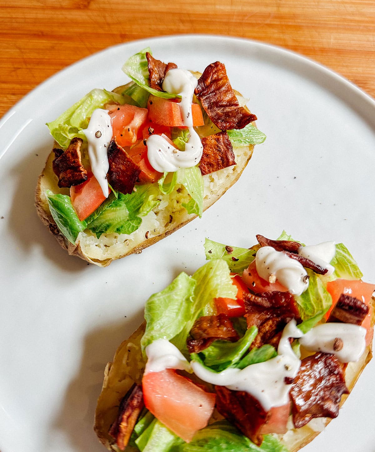 Two baked potato halves topped with lettuce, tomato, bacon pieces, and a drizzle of white sauce on a white plate.