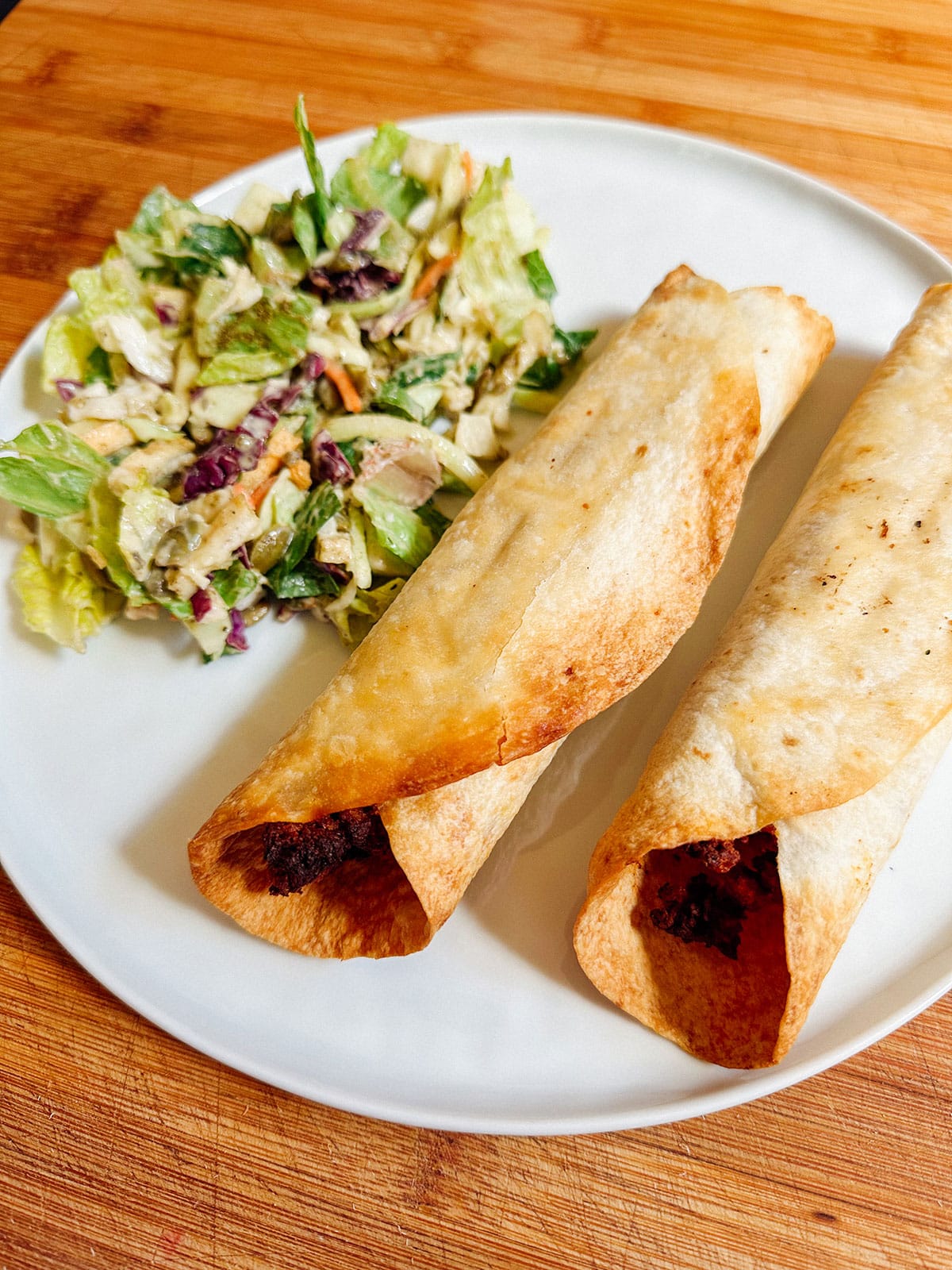 Two baked taquitos with a dark filling are served on a white plate next to a small portion of chopped salad with dressing.