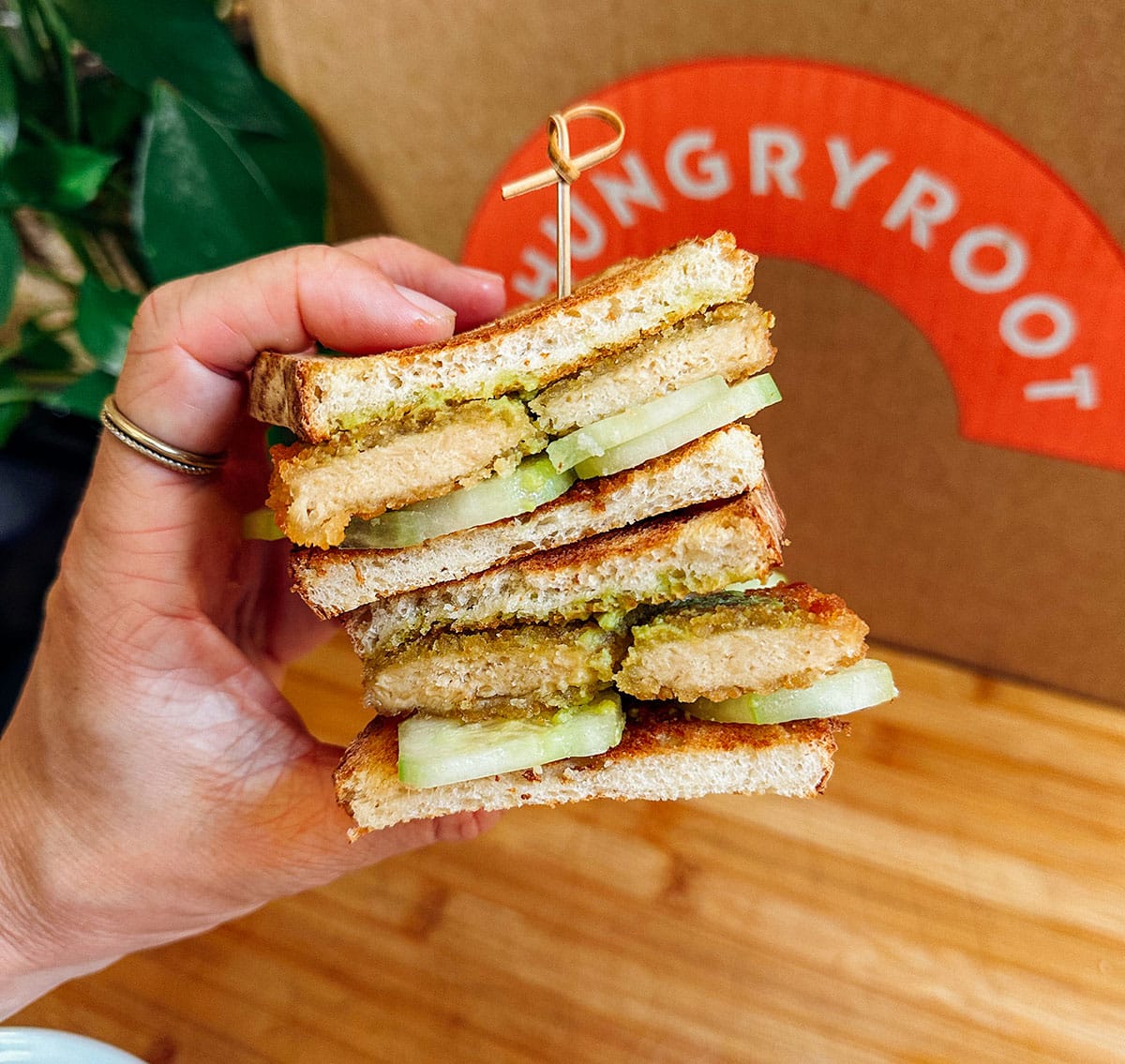 A hand holds a stacked sandwich with breaded patties, sliced cucumber, and sauce between toasted bread, in front of a box labeled Hungryroot.