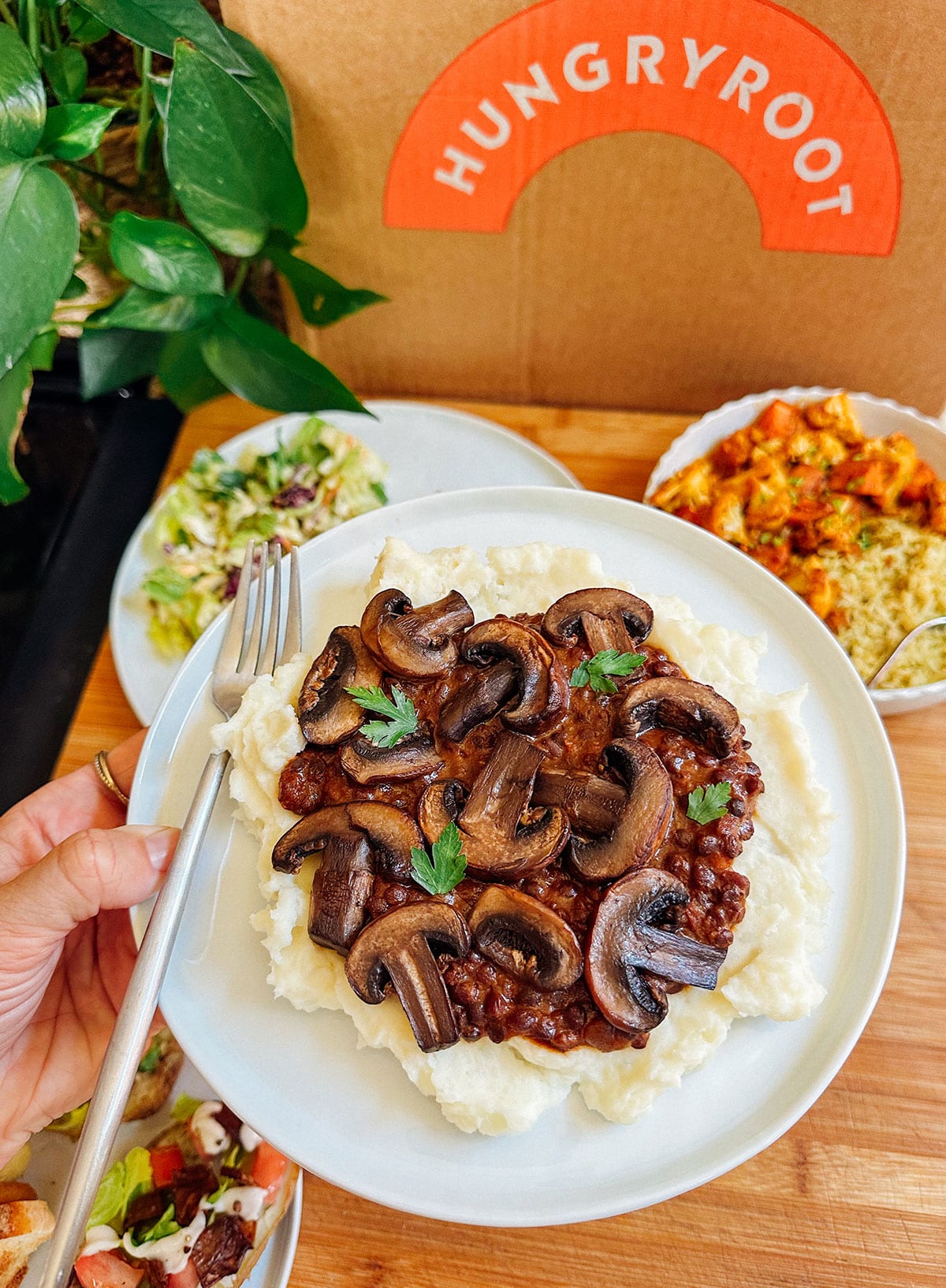 A plate of mashed potatoes topped with mushrooms and parsley, held above a table with other dishes, in front of a Hungryroot box.