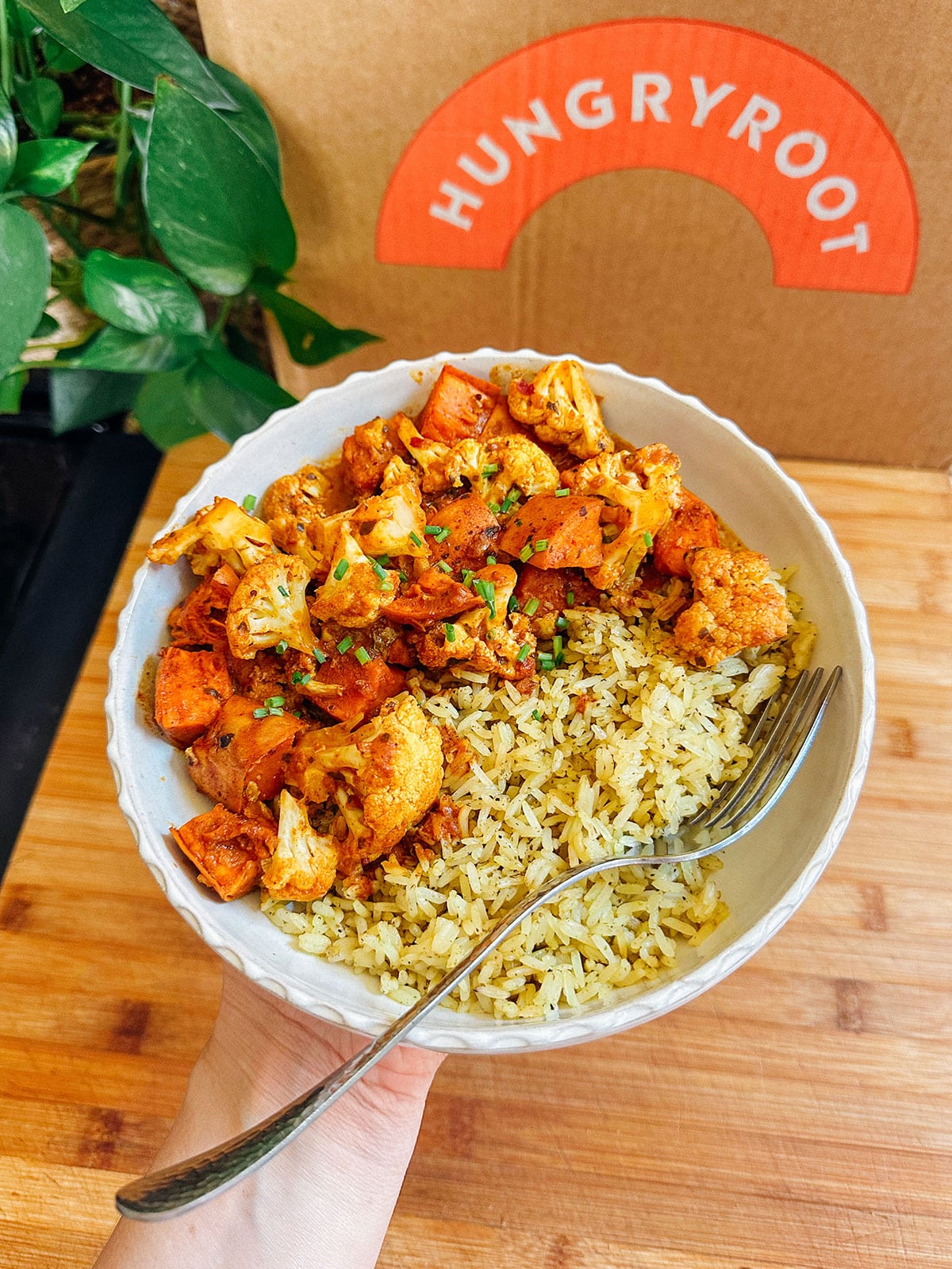 A hand holds a bowl of yellow rice and seasoned roasted vegetables with a fork, in front of a Hungryroot box on a wooden surface.