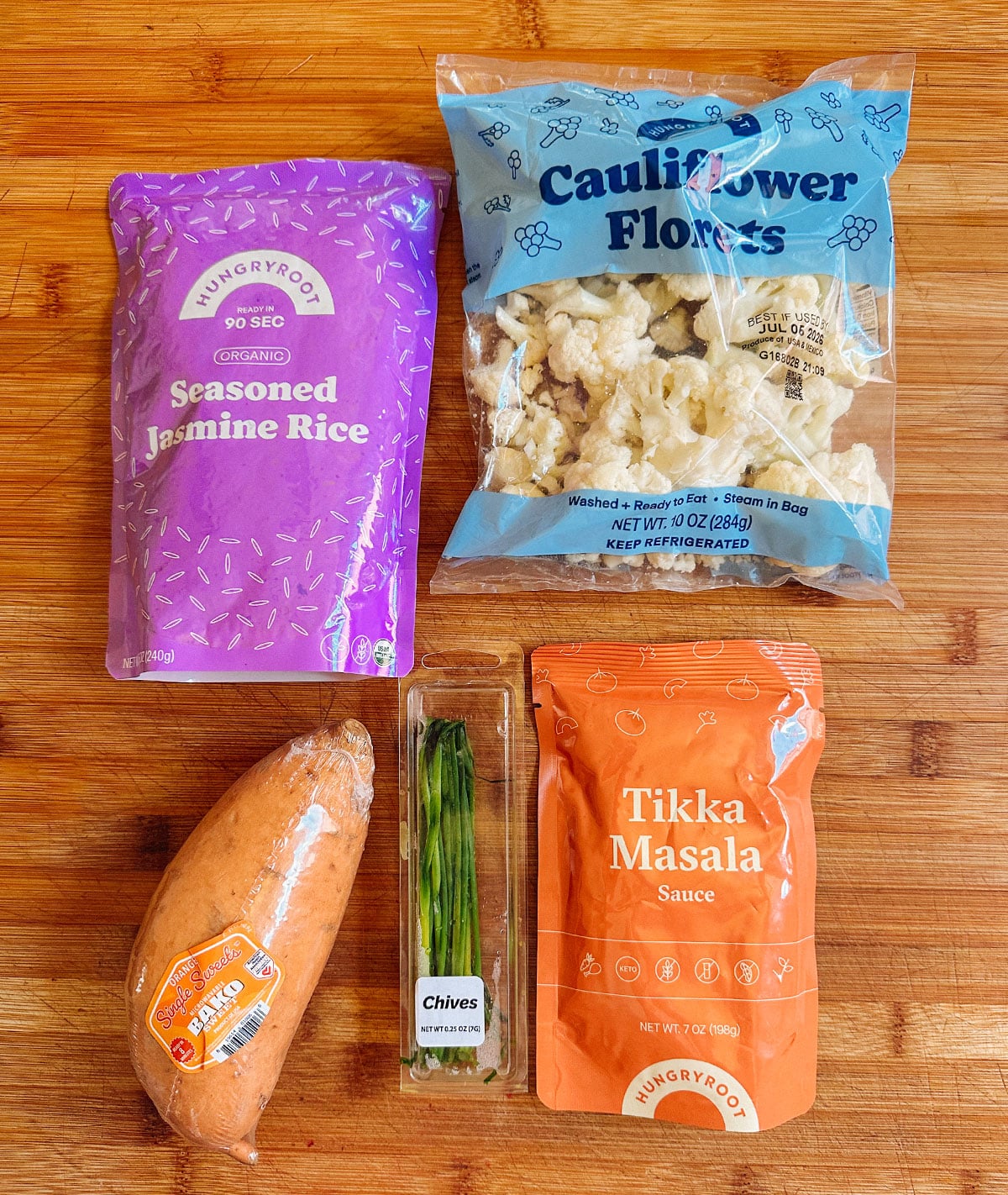 Four packaged food items—seasoned jasmine rice, cauliflower florets, tikka masala sauce, and fresh chives—plus one raw sweet potato displayed on a wooden surface.