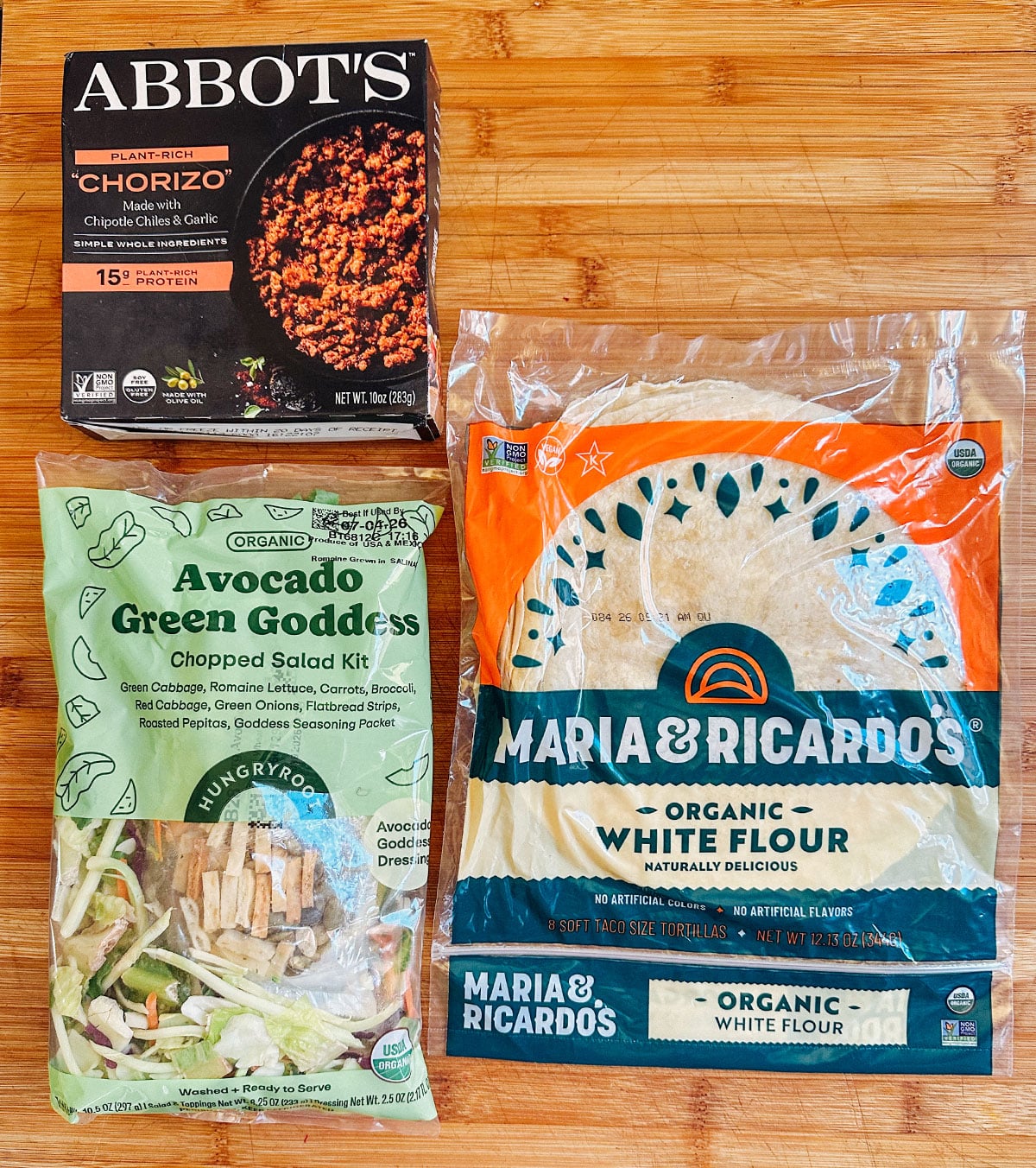 Three food items on a wooden surface: Abbotts chorizo, an Avocado Green Goddess chopped salad kit, and Maria & Ricardos organic white flour tortillas.