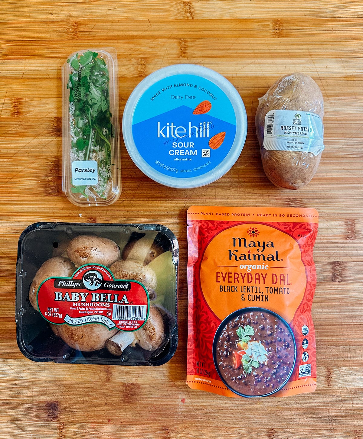 Five ingredients on a wooden surface: parsley, dairy-free sour cream, a russet potato, baby bella mushrooms, and a pouch of black lentil dal.