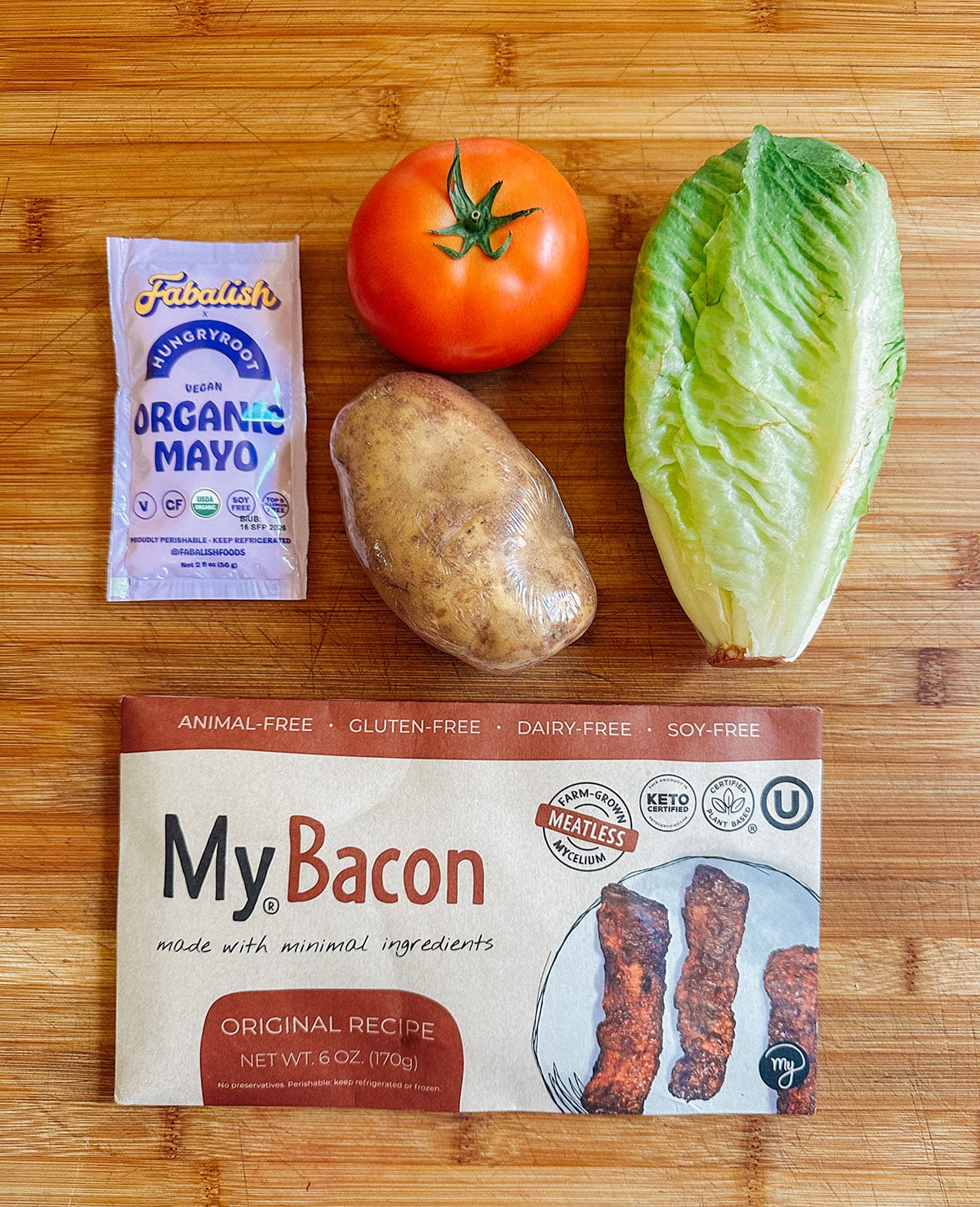 A package of vegan mayo, a tomato, a potato, a romaine lettuce leaf, and a box of plant-based bacon arranged on a wooden cutting board.