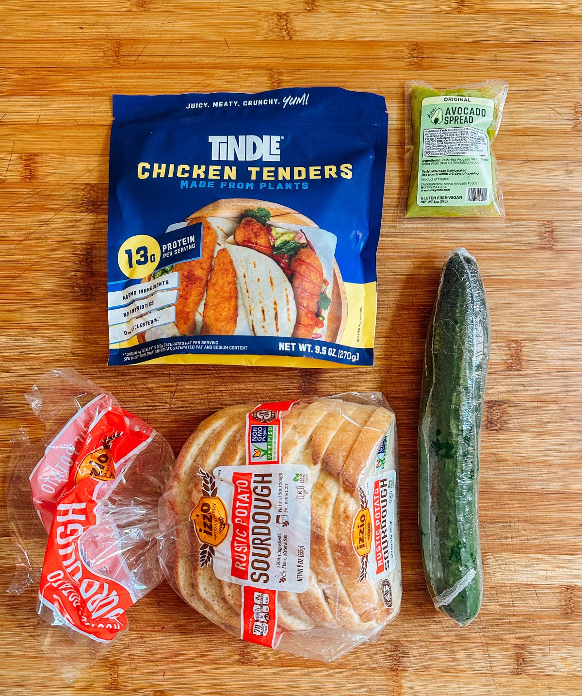 A package of plant-based chicken tenders, avocado spread, a cucumber, and a bag of sliced sourdough bread on a wooden surface.