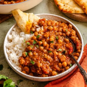 A bowl of white rice topped with chickpea curry, garnished with herbs, served with naan bread; a spoon rests in the bowl.