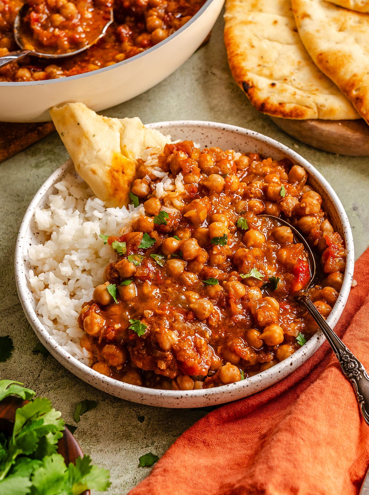 A bowl of white rice topped with chickpea curry, garnished with herbs, served with naan bread; a spoon rests in the bowl.