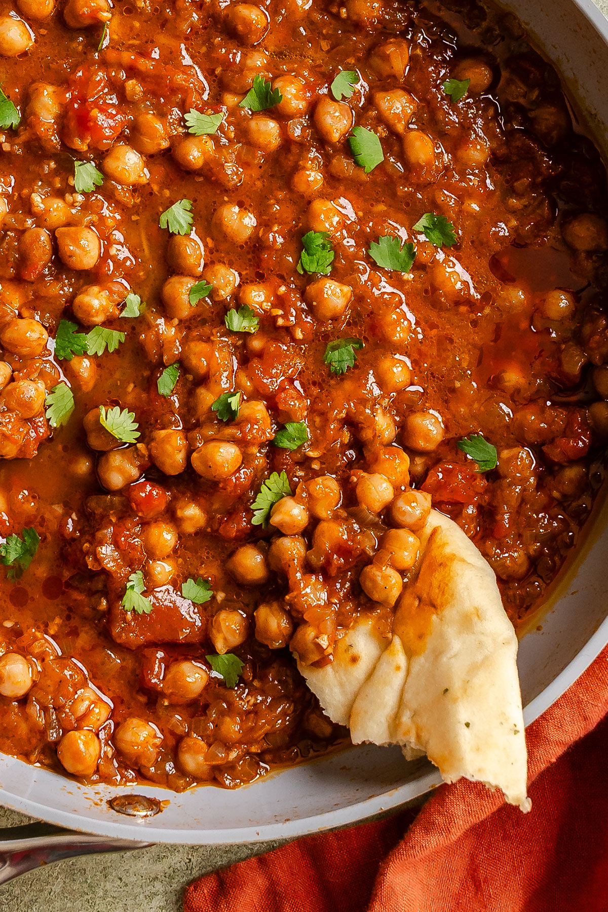 Chickpea curry with tomato sauce, garnished with cilantro, served in a pan with a piece of naan bread dipping in the curry.