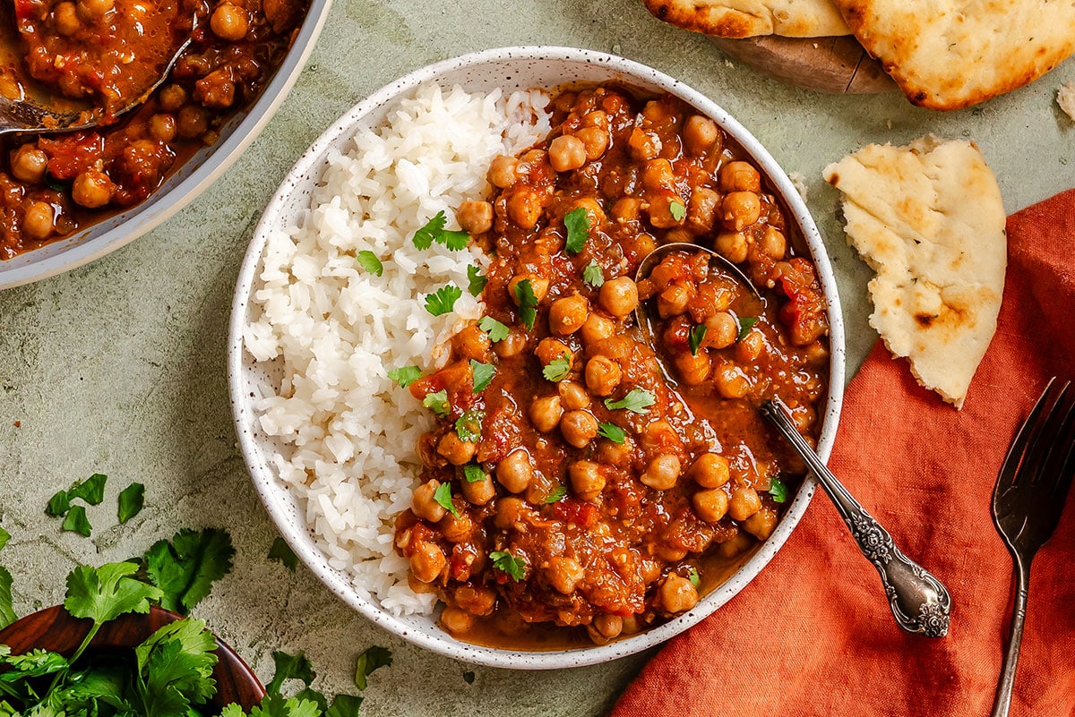 A bowl with white rice and chickpea curry garnished with cilantro, served with naan bread on the side and a spoon in the bowl.