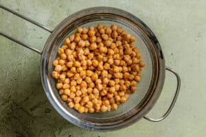 A metal strainer filled with cooked chickpeas is placed on a light green textured surface.