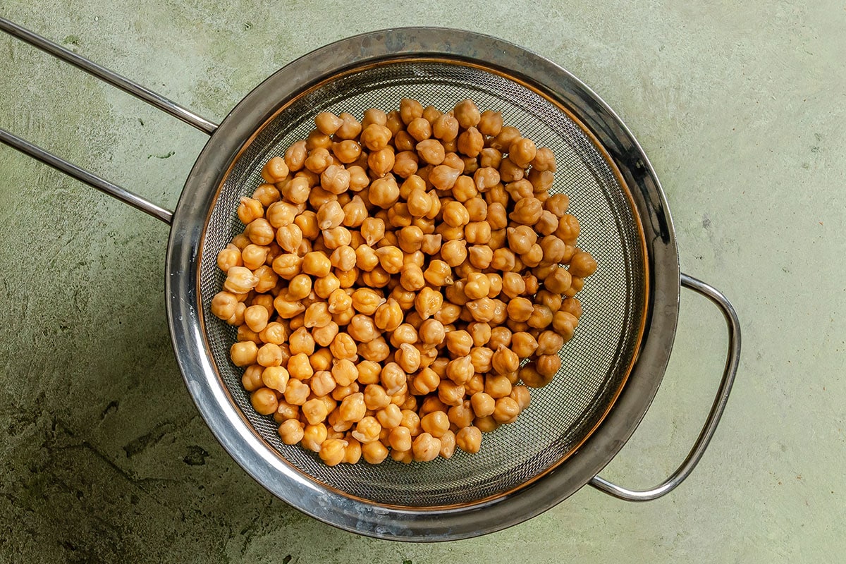 A metal strainer filled with cooked chickpeas is placed on a light green textured surface.