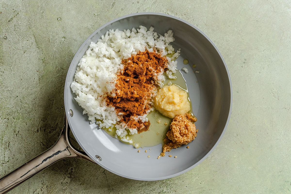 A frying pan containing chopped onion, ground spices, ginger paste, and minced garlic on a light green surface.