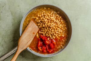 A skillet with chickpeas, chopped tomatoes, diced onions, and broth being stirred with a wooden spatula on a light green surface.