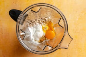 A blender contains oats, yogurt, an egg, and a splash of milk, viewed from above on a yellow surface.
