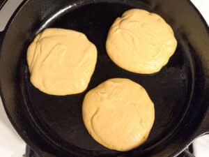 Three uncooked pancakes are being cooked on a black cast iron skillet.