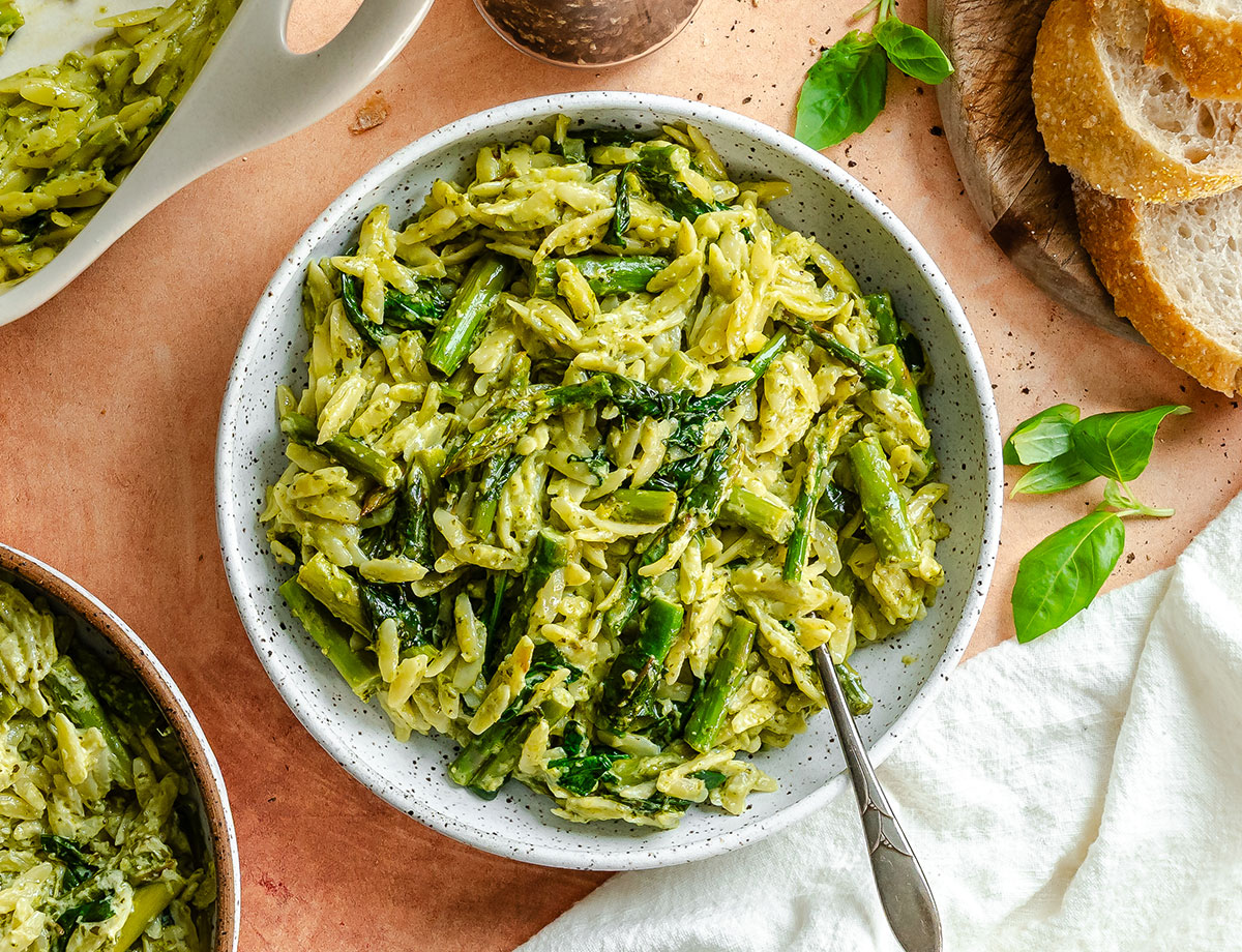 Bowl of orzo pasta mixed with green vegetables and herbs, beside slices of bread, basil leaves, and a serving dish on a wooden surface.