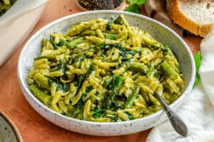 A speckled bowl filled with orzo pasta, green vegetables, and a creamy sauce, with a spoon resting inside the bowl. Slices of bread are visible in the background.
