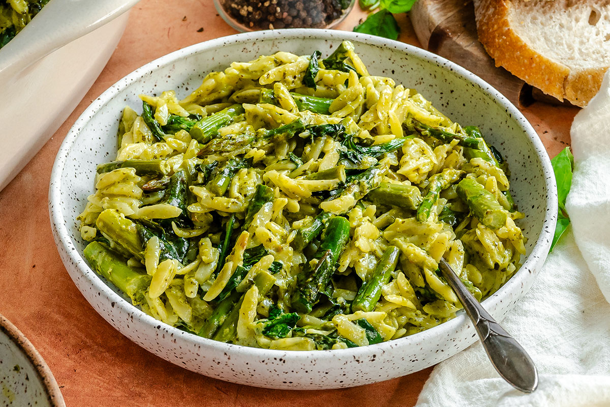 A speckled bowl filled with orzo pasta, green vegetables, and a creamy sauce, with a spoon resting inside the bowl. Slices of bread are visible in the background.