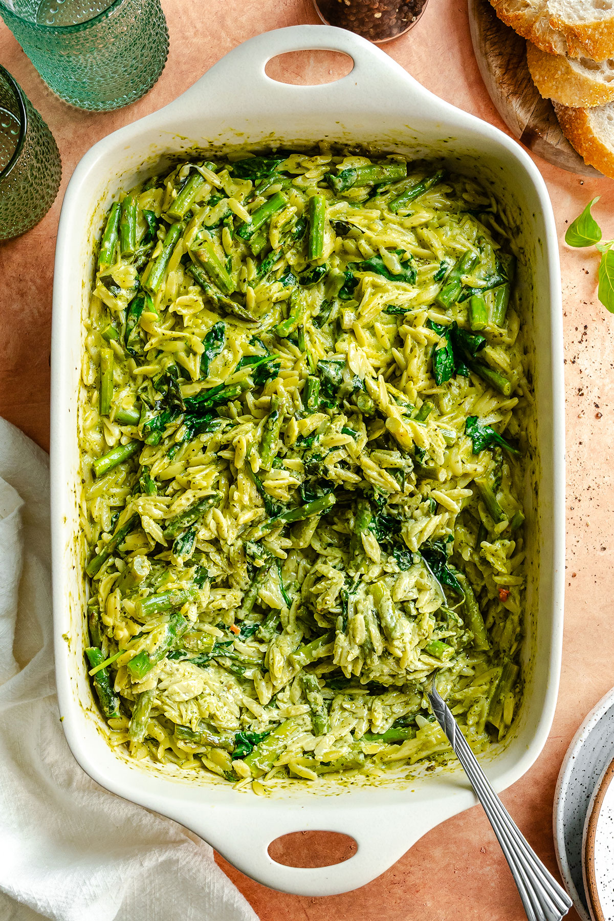 Rectangular baking dish filled with creamy green pesto orzo pasta mixed with spinach and asparagus, with a serving spoon on the side.