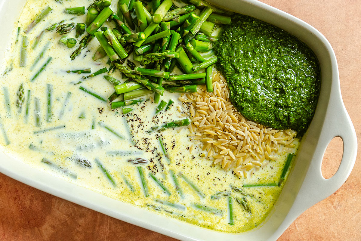 Uncooked casserole in a white dish with milk, chopped asparagus, green pesto, orzo pasta, and seasonings arranged separately.