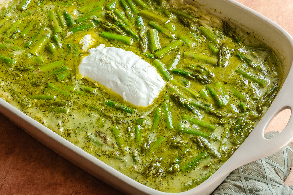 A casserole dish filled with baked asparagus in a green sauce, topped with a dollop of white cream, sits on a brown surface next to an oven mitt.