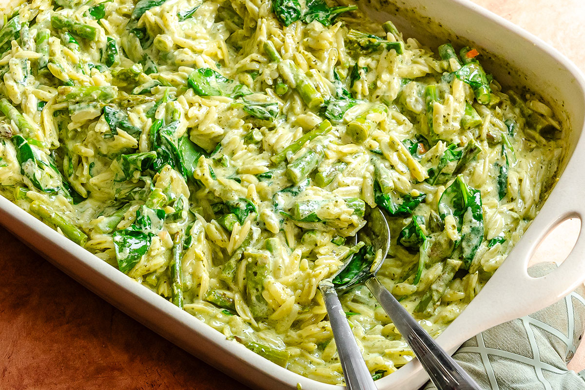 A baking dish filled with creamy orzo pasta mixed with spinach and cheese, with a serving spoon and tongs resting inside.