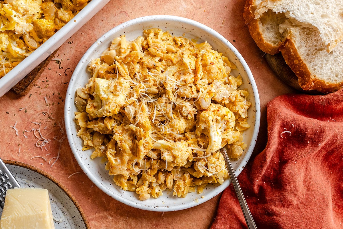 A bowl of macaroni and cheese topped with shredded cheese sits on a plate, next to slices of bread on a wooden surface with a cloth napkin and a gold fork.