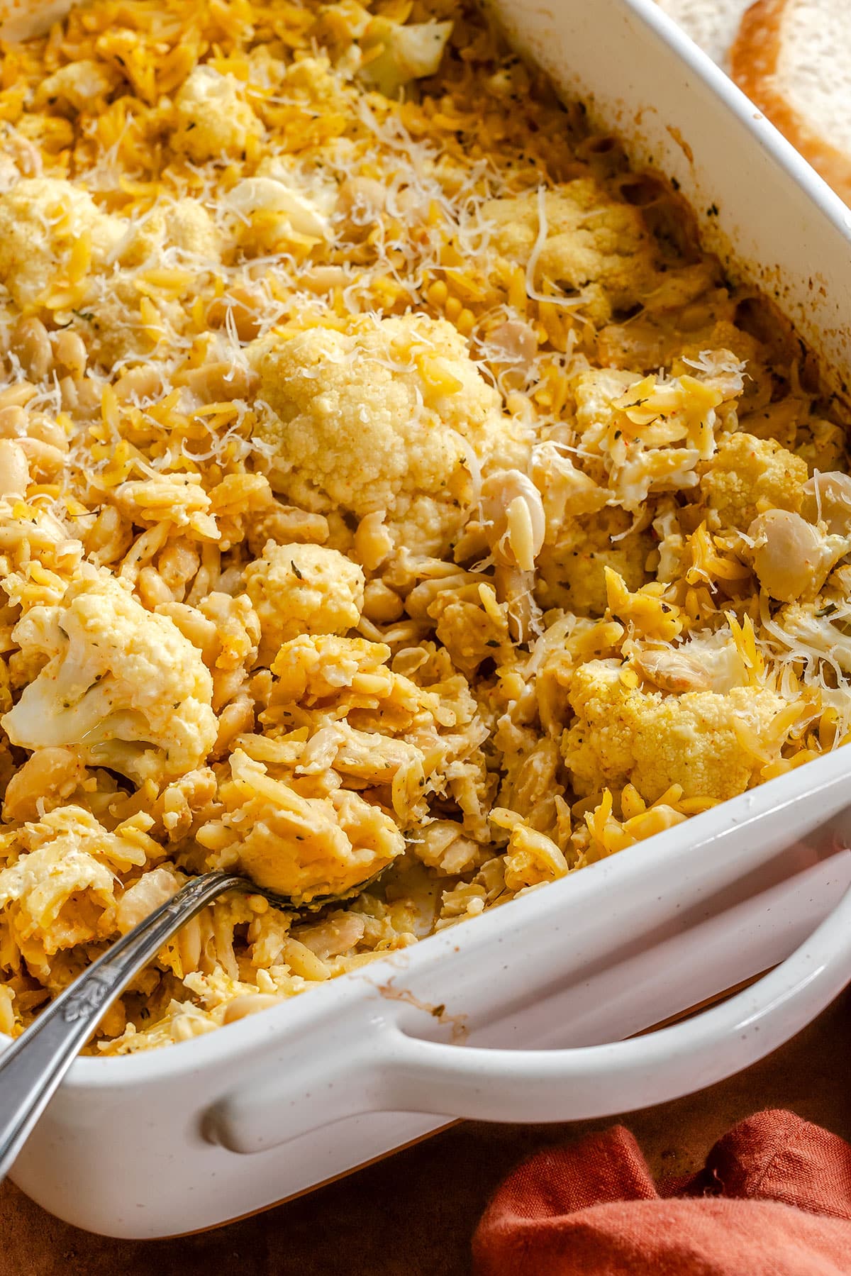 A close-up of a white baking dish filled with cheesy cauliflower and pasta casserole, with a serving spoon partially scooping some out.