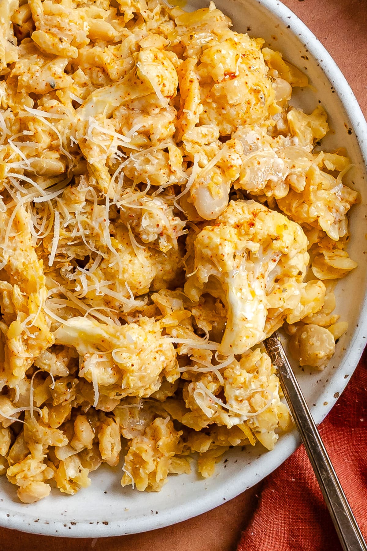Close-up of a plate with cauliflower, white beans, shredded cheese, and pasta, mixed together in a creamy sauce, with a fork resting on the side.