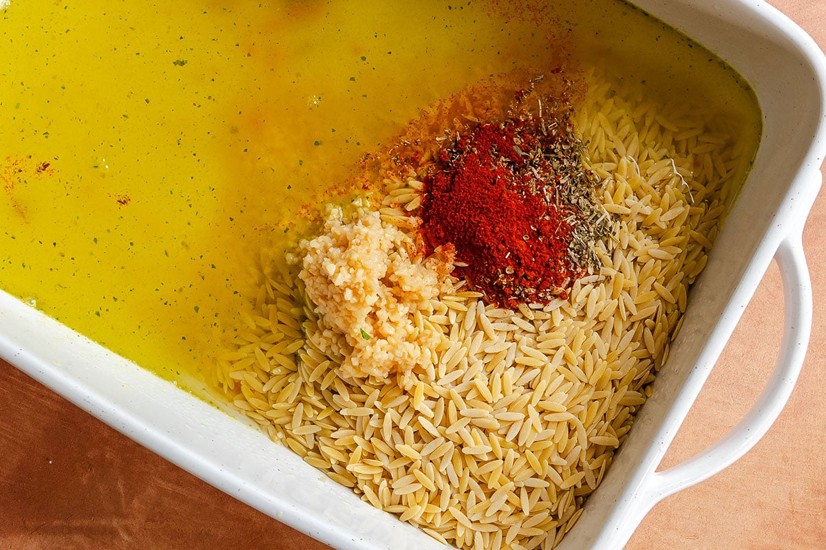 Uncooked orzo, chopped garlic, spices, and herbs are in a white baking dish filled with yellow broth, ready for mixing.