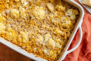 Baked casserole with shredded cheese, cauliflower florets, white beans, orzo pasta, and grated cheese on top in a white baking dish.