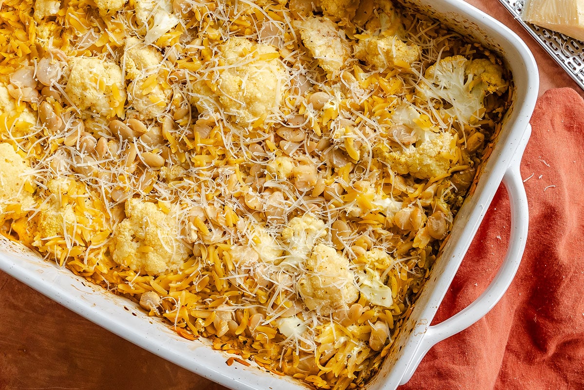 Baked casserole with shredded cheese, cauliflower florets, white beans, orzo pasta, and grated cheese on top in a white baking dish.