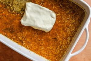 Rectangular white baking dish with uncooked orzo pasta, orange-colored seasoned liquid, and a block of white cheese placed in the center.
