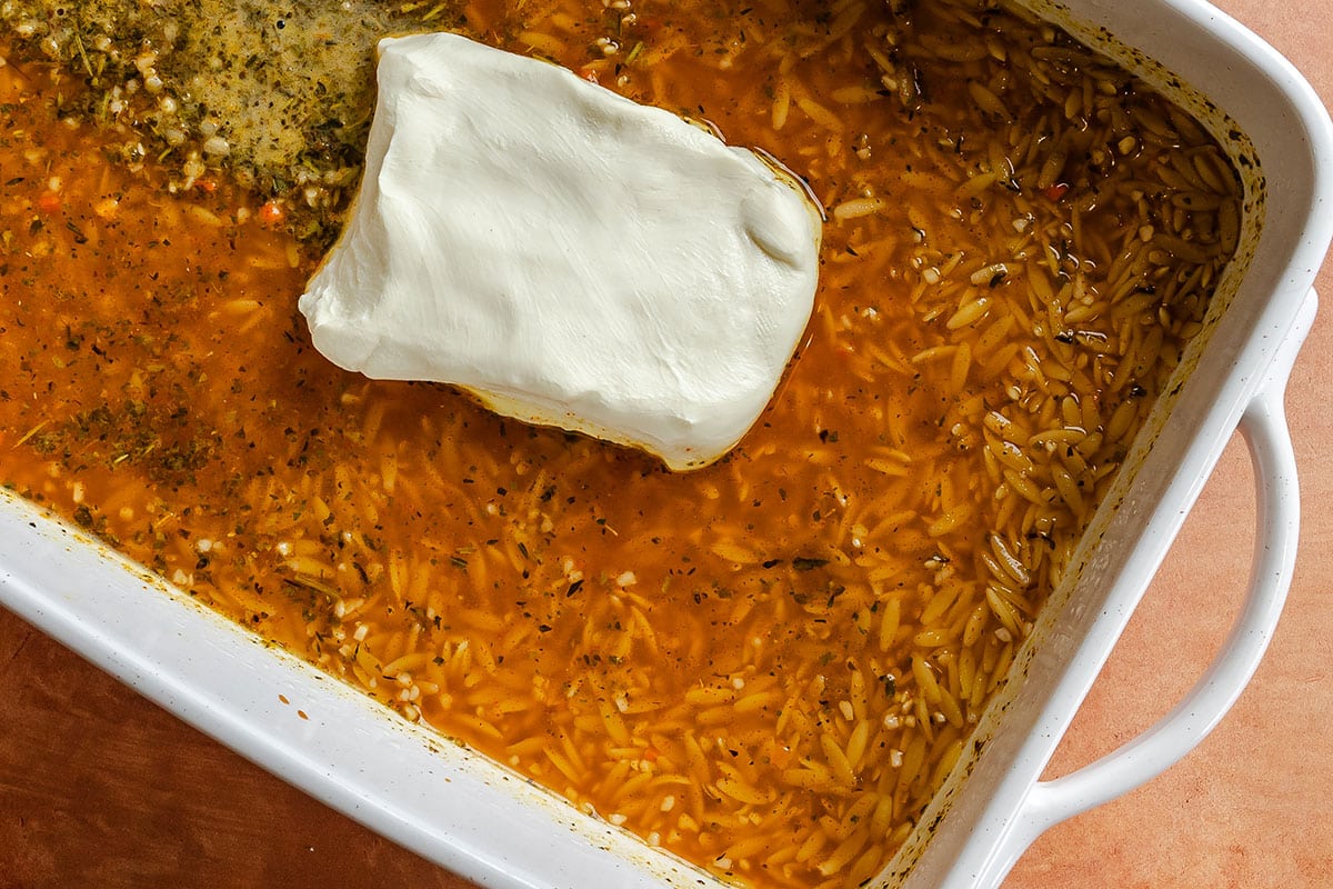 Rectangular white baking dish with uncooked orzo pasta, orange-colored seasoned liquid, and a block of white cheese placed in the center.