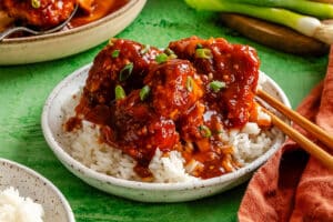 A bowl of white rice topped with saucy meatballs and chopped green onions, with chopsticks resting on the side.