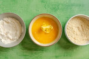 Three bowls on a green surface: one with flour, one with beaten eggs and butter, and one with breadcrumbs.