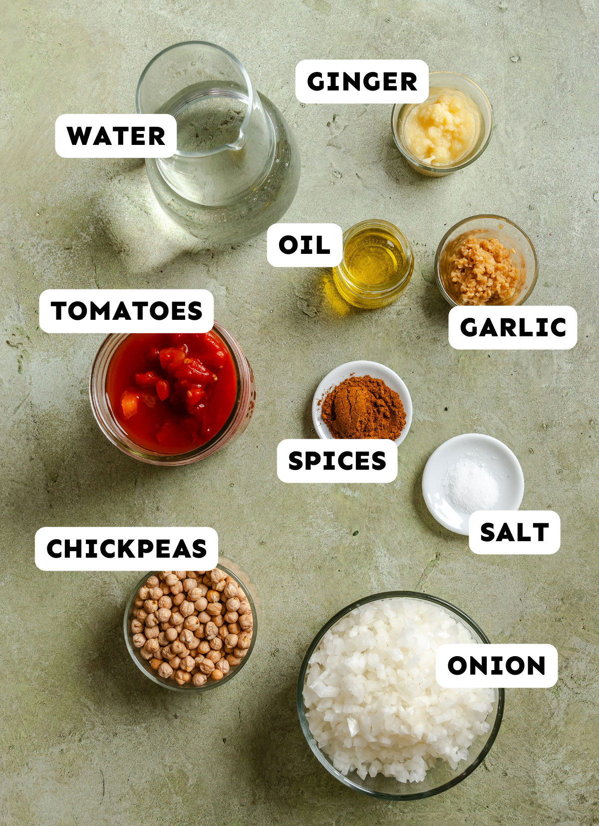 Overhead view of ingredients in bowls: water, ginger, oil, garlic, salt, spices, tomatoes, chickpeas, and chopped onion—perfectly arranged on a textured surface—showcases the essentials for an authentic Chana Masala chickpea recipe.