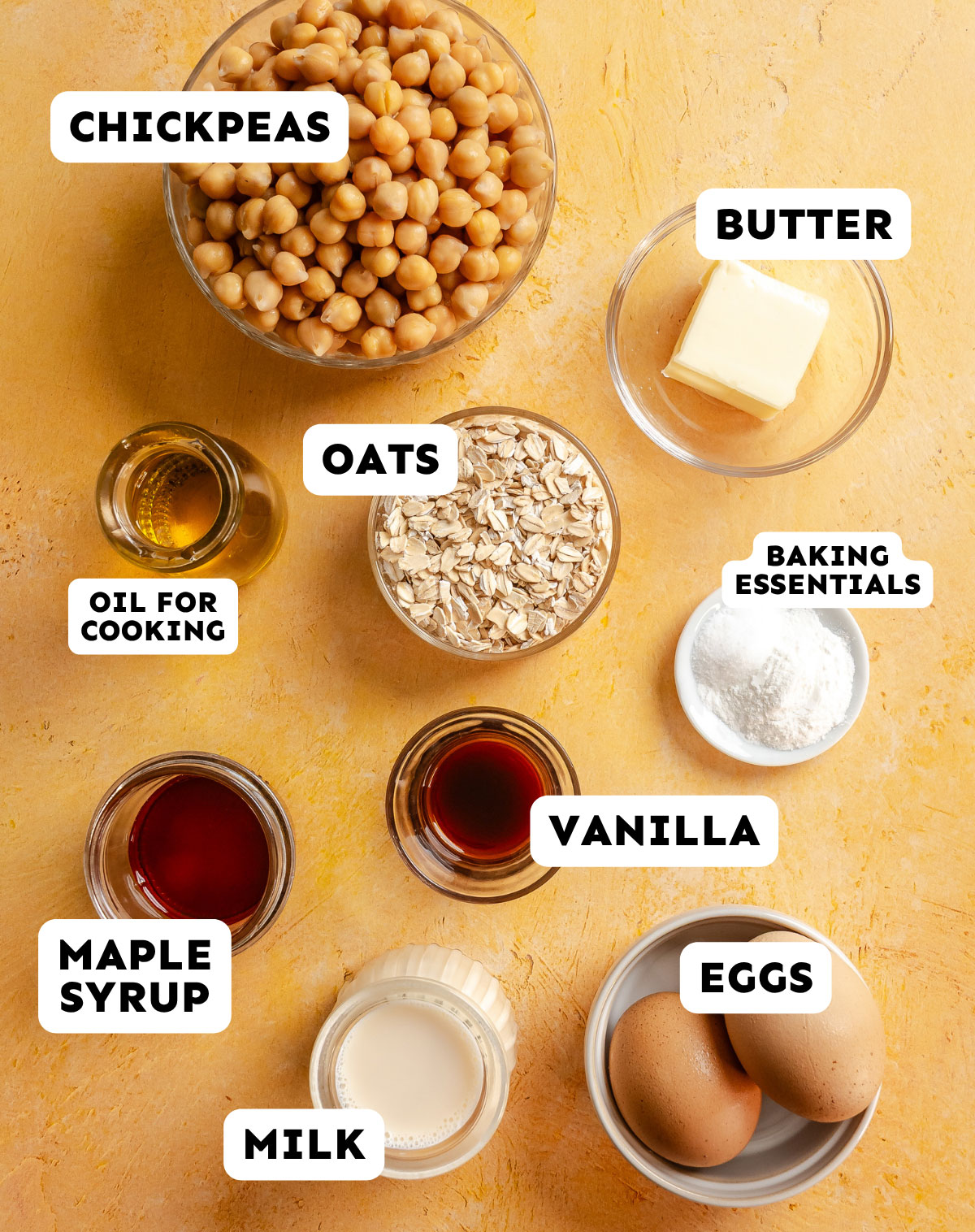 A top-down view of various baking ingredients on a yellow surface, including chickpeas, butter, oats, oil, baking powder, maple syrup, vanilla, milk, and eggs, each labeled—perfect for making delicious Chickpea Flour Pancakes.