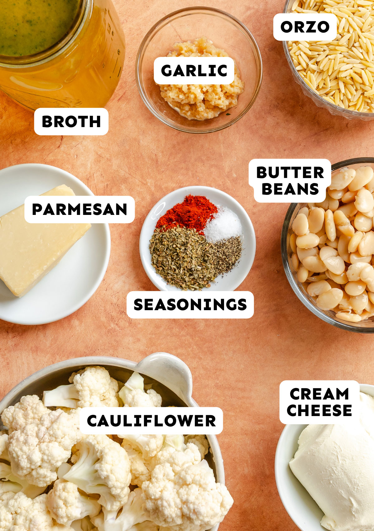 Ingredients on a countertop: jar of broth, minced garlic, orzo pasta, butter beans, cream cheese, cauliflower florets, Parmesan cheese, and a small plate with mixed seasonings—everything you need for an easy Dump and Bake Cauliflower Casserole or a creamy Parmesan Orzo dish.