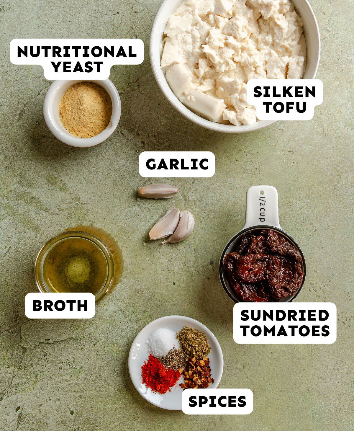 Six labeled ingredients for a recipe are shown: nutritional yeast, silken tofu, garlic, broth, sundried tomatoes, and spices, all arranged on a green surface.