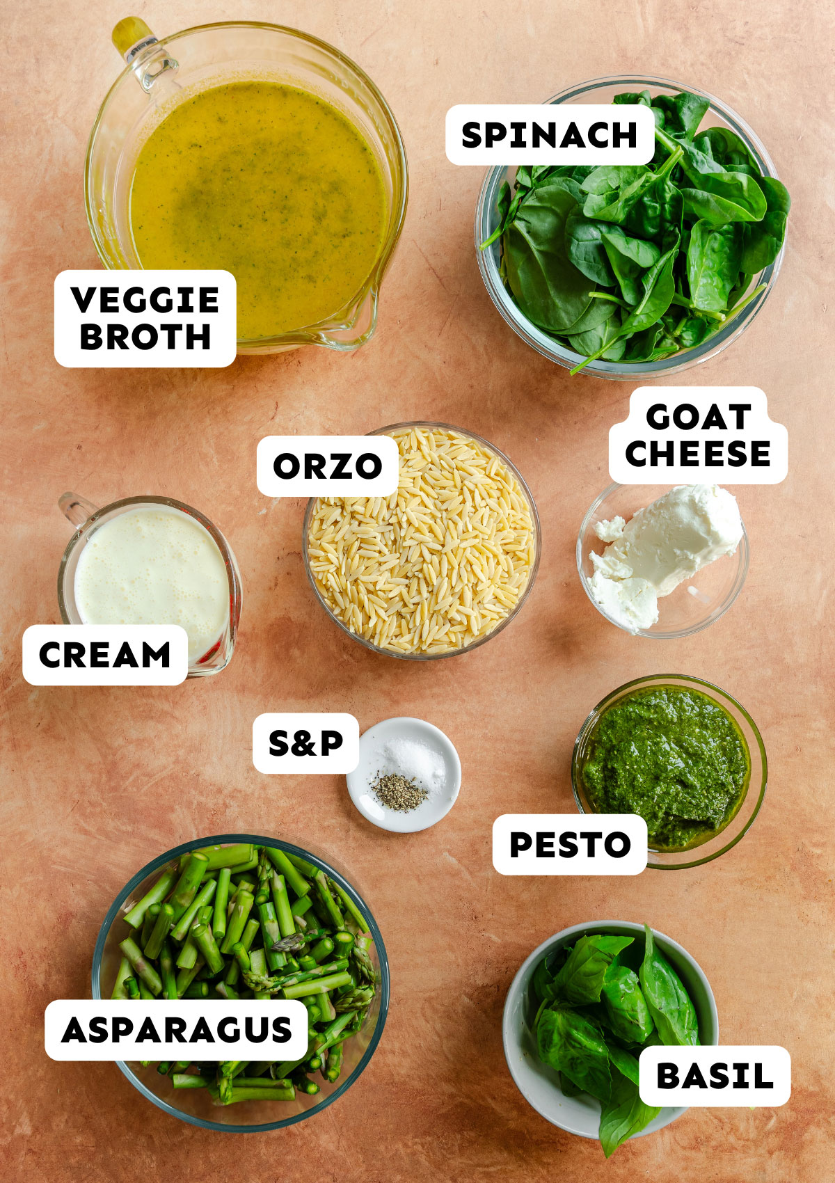 Ingredients for a pasta dish arranged on a countertop: veggie broth, spinach, orzo, goat cheese, cream, salt and pepper, pesto, asparagus, and basil.