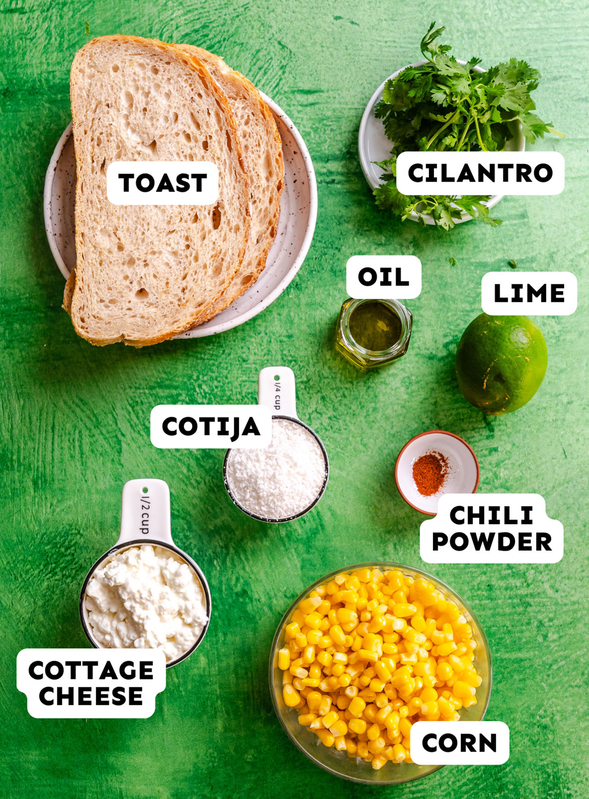 Slices of toast, cilantro, oil, lime, chili powder, corn, cottage cheese, and cotija cheese are arranged on a green background for a vibrant take on Mexican street corn toast.