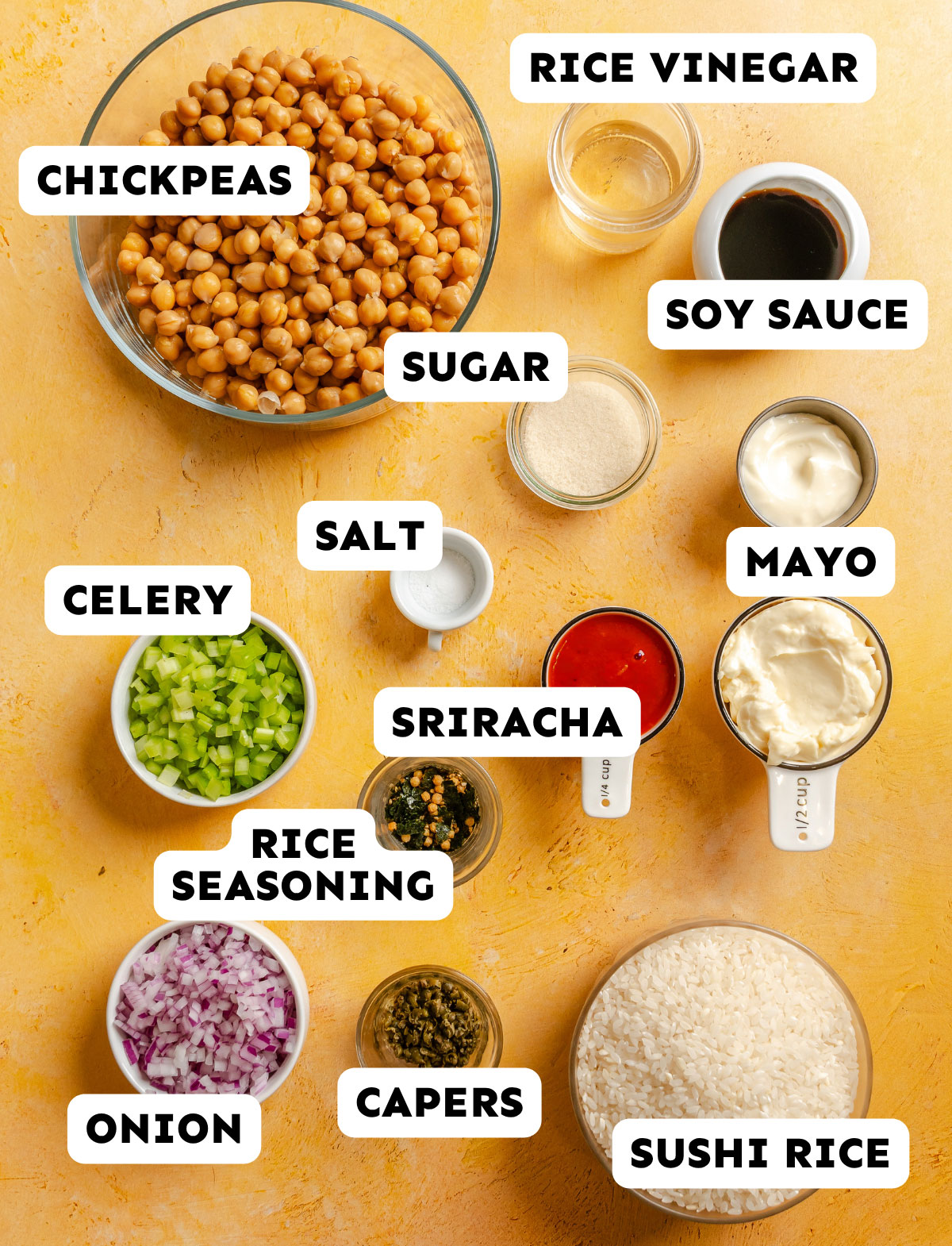 Ingredients for a chickpea sushi bowl are arranged on a yellow surface, each labeled: chickpeas, rice vinegar, soy sauce, sugar, salt, mayo, celery, sriracha, rice seasoning, onion, capers, and sushi rice.