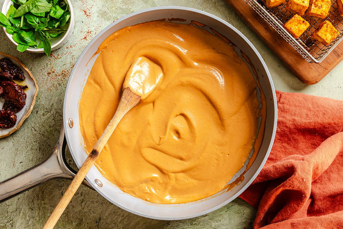 A wooden spoon rests in a pan filled with creamy orange sauce, with fresh basil, sun-dried tomatoes, and tofu cubes nearby on a textured surface.