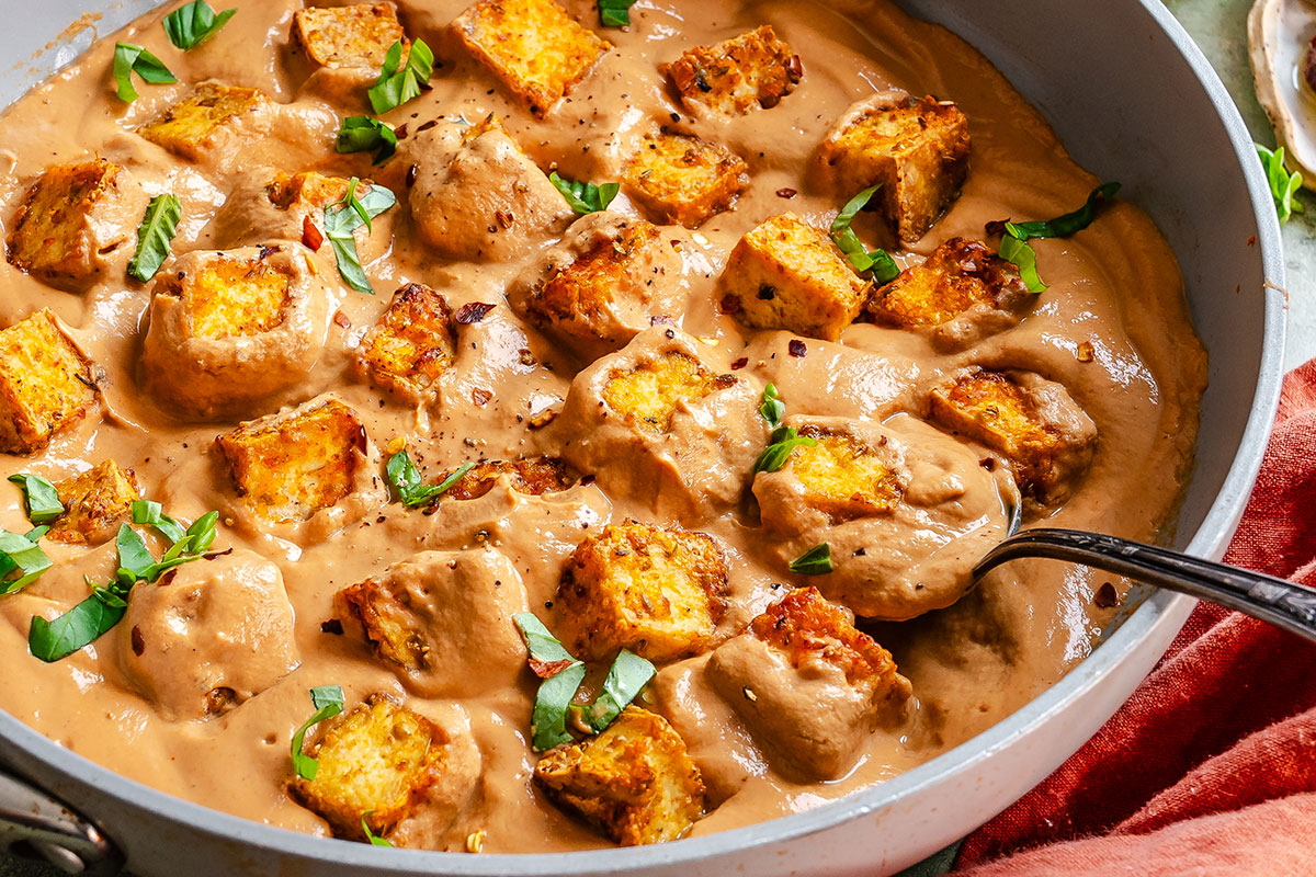 A skillet filled with creamy brown sauce, crispy bread cubes, and chopped fresh herbs, with a spoon partially submerged.