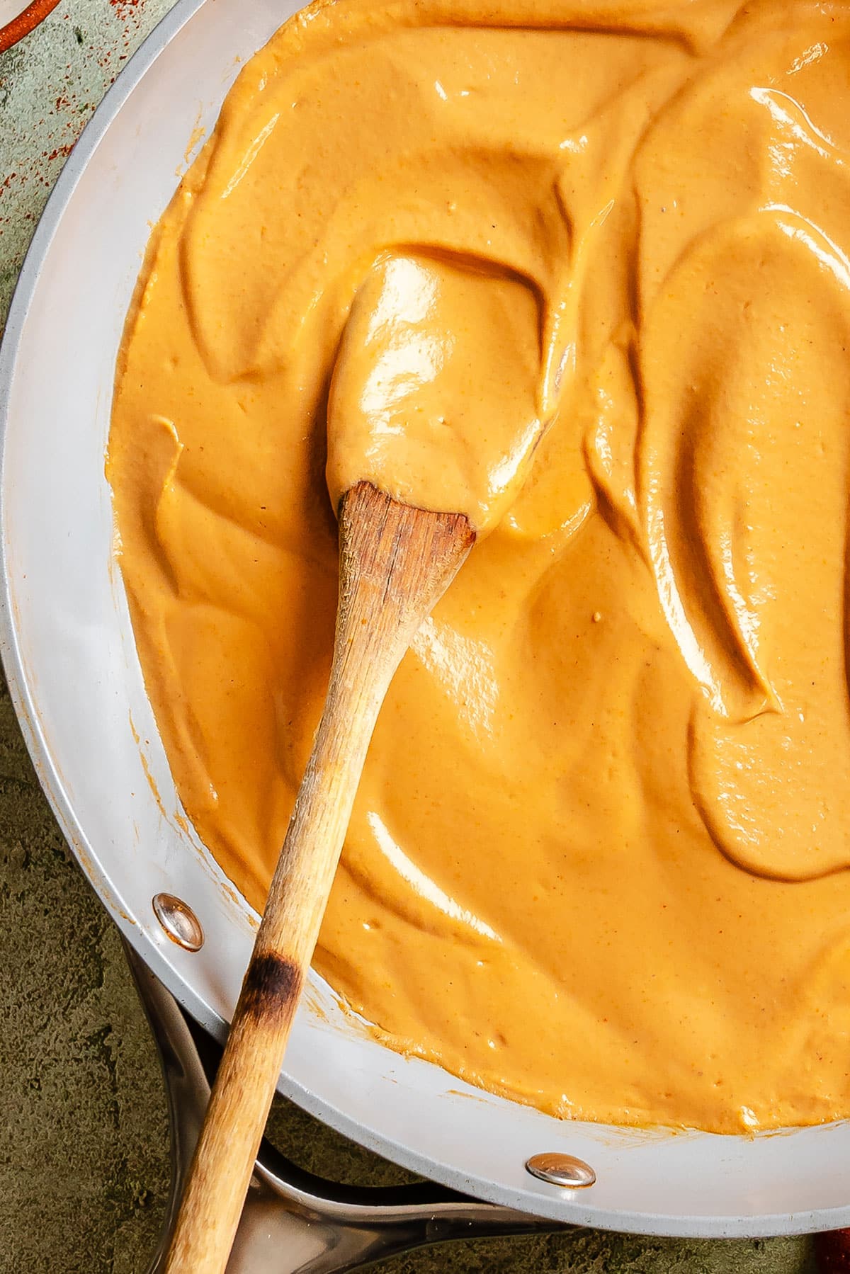 A wooden spoon stirs a thick, creamy orange sauce in a metal pan.