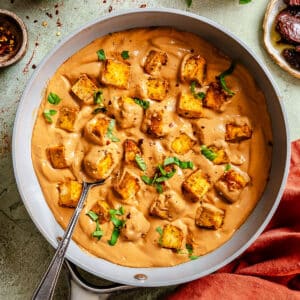 Pan of creamy sauce with golden tofu cubes, garnished with chopped herbs. A spoon rests in the pan, surrounded by small bowls of spices and fresh greens.